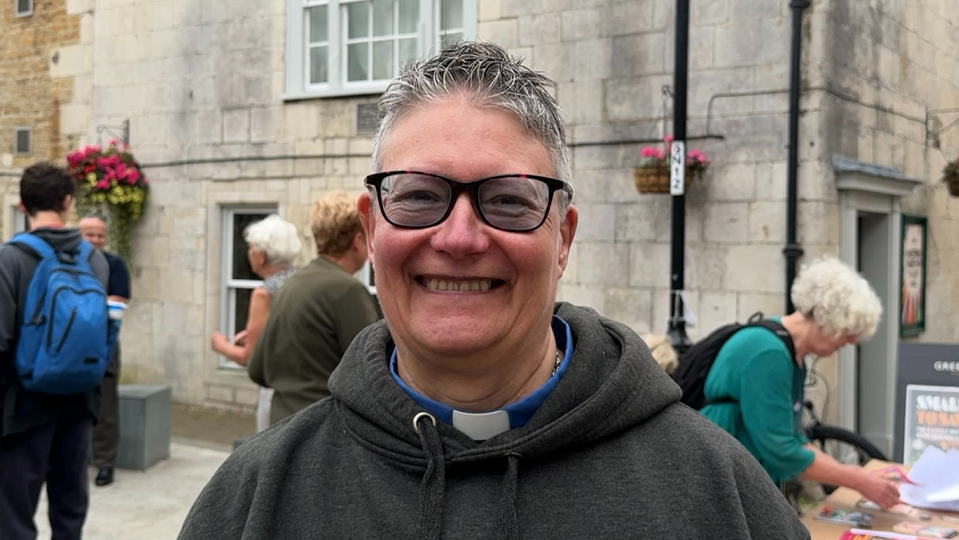 Angie McLachlan has grey/white hair that has been gelled up, a grey hooded top and a blue shirt with a dog collar. She is also wearing black-rimmed glasses 