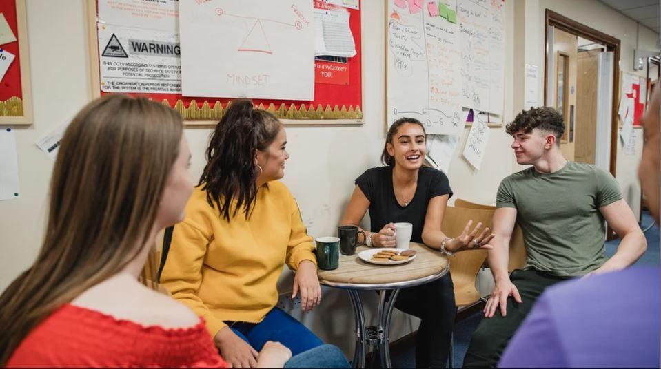 A group of teenagers sit around a wooden table. There are mugs and biscuits on the table and posters and notes written on the walls.