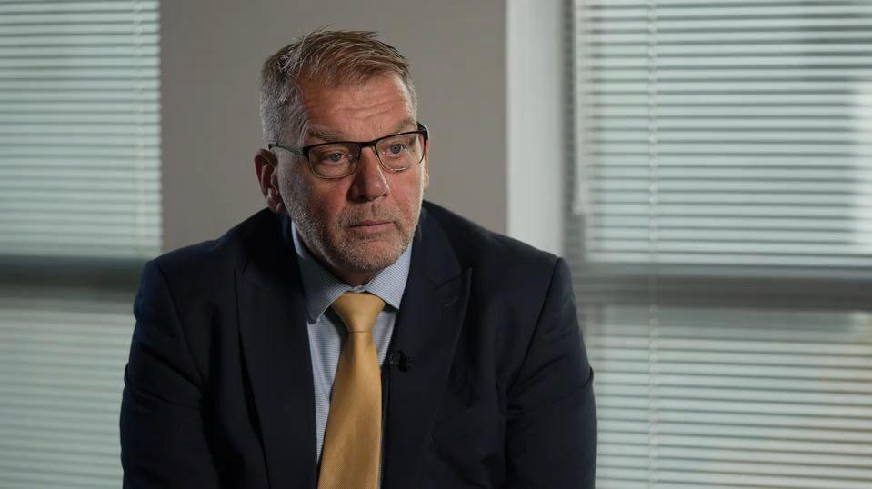 Gary Haskins sits in an office. He wears a navy blue suit jacket, light blue shirt and a gold tie. He has mousey brown hair and black glasses.