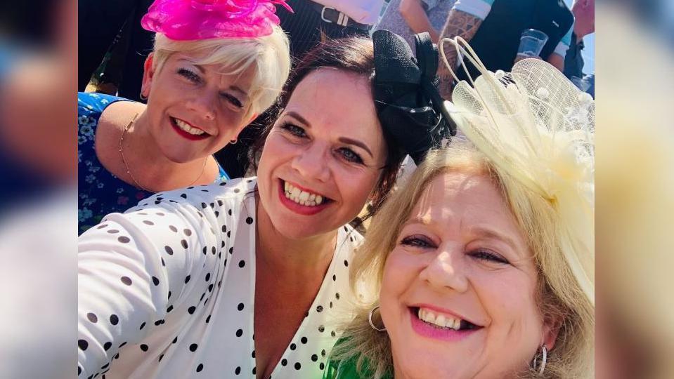 Three women looking into the camera taking a selfie. The woman on the left has blonde short hair and a pink headpiece and a dark blue dress with light blue flowers. She is wearing a gold necklace. The woman in the middle has long brown hair tied up and a black headpiece. She is wearing a V-neck white and black spotty dress. The woman on the right of the photo has blonde hair and has a yellow headpiece on. She has silver hoops and is wearing a green outfit. 