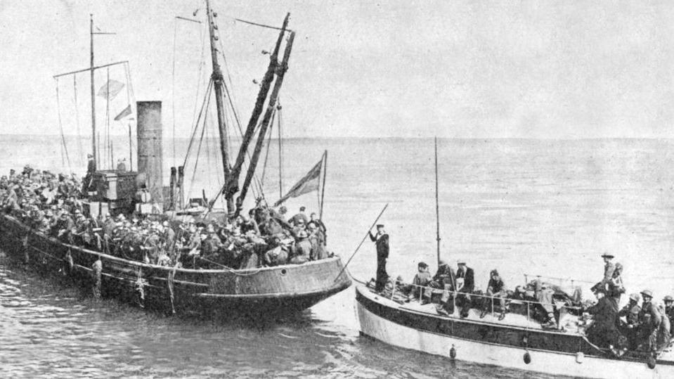 A black and white archive image of a little boat in open water meeting another vessel packed with troops.