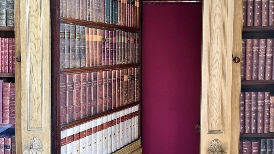 The door of the secret bookshelf has been opened to show that there is a door behind it that leads to another room