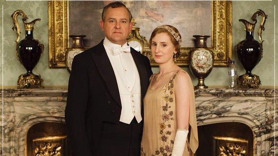 The Early of Grantham and Lady Edith standing in front of a shelf with a bottle on