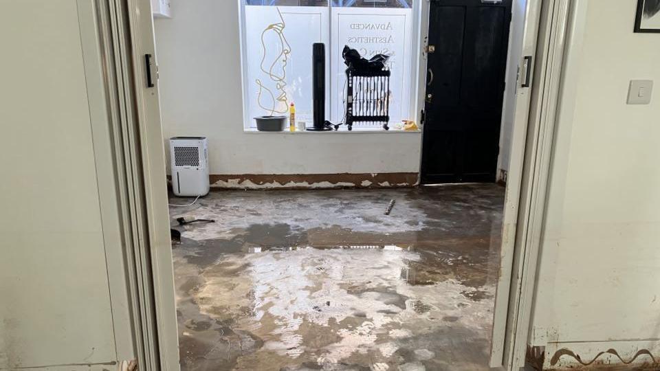 An internal photo of a shop comprising of a doorway between two internal rooms. The concrete floor is wet and walls appear damaged, with paintwork stripped away. The front window can be seen, with artwork depicting the outline of a woman's face. Items on the windowsill include a bowl, bottle of bleach and portable radiator. A dehumidifieris on the floor.