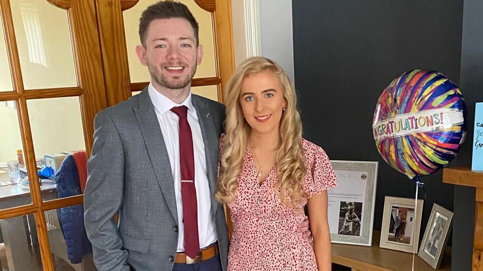 Cahir wearing grey suit and red tie beside Shannon, wearing pink dress, with long blonde hair. They are standing beside family photos and a congratulations balloon