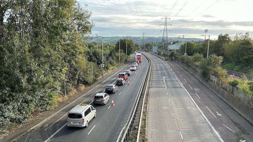 A line of cars sitting in traffic on a motorway bypass which has one lane closed with orange and white cones. 
The sky is grey with clouds and the road is lined with green shrubs and trees. 
The other lane is empty.