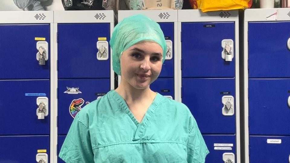 Tabatha wears surgeon scrubs and a cloth hat in a green colour. She stands in front of blue lockers.