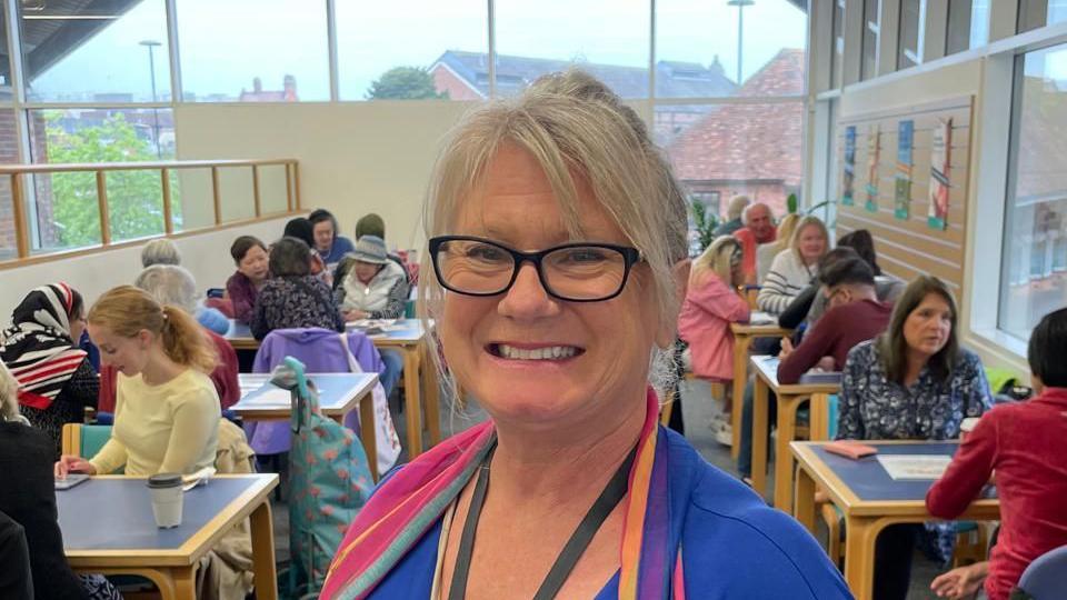 An older woman with blonde hair wearing glasses from the shoulders up. In the background people are sat at tables.