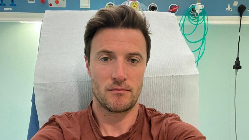 A man in a brown T-shirt takes a selfie in a hospital bed. He is not smiling.