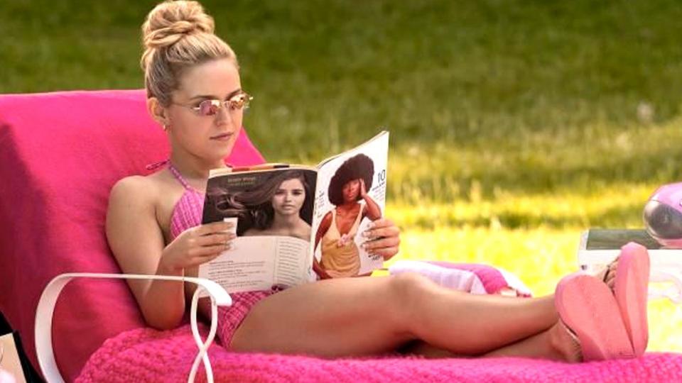 A woman in a pink bikini lies on a deck chair covered in pink blankets, reads a magazine. there are pink towels, a tote bag and a radio next to her.