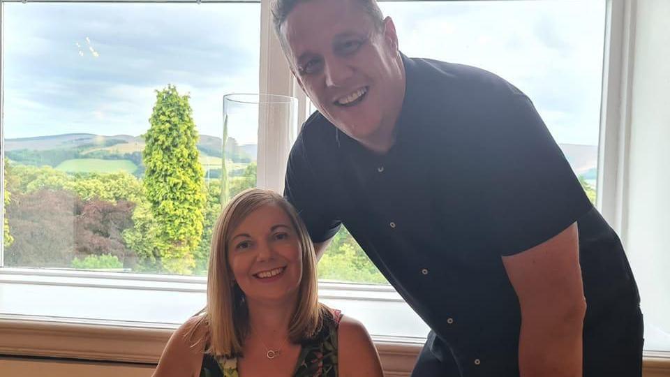 A woman, Anna, sits down next to a man, Gary. Anna has shoulder length blonde hair and wears a tropical patterned top. Gary has a dark shirt on. They are both smiling into the camera.