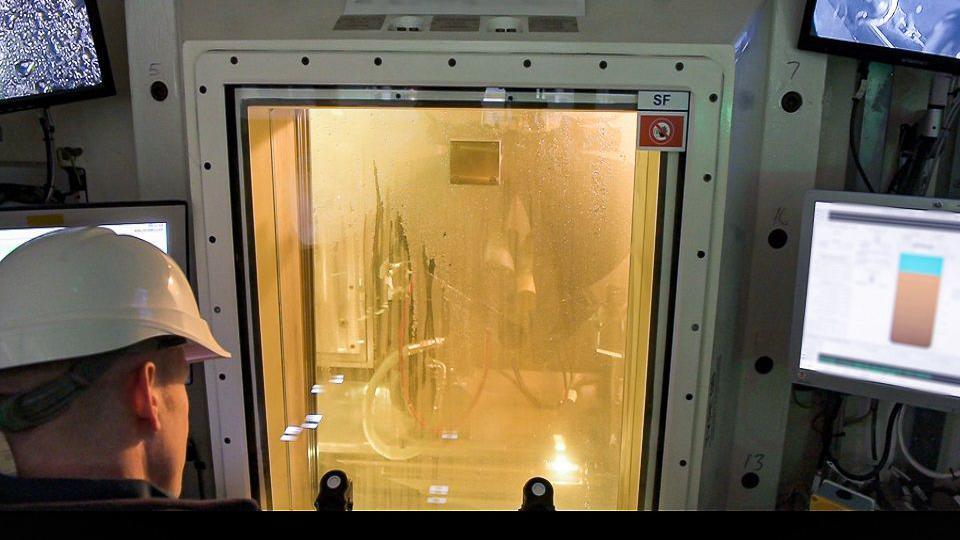 A man wearing a white hard-hat looks through the window of a container holding a complicated piece of machinery at Sellafield. There are small monitor screens either side of the container.