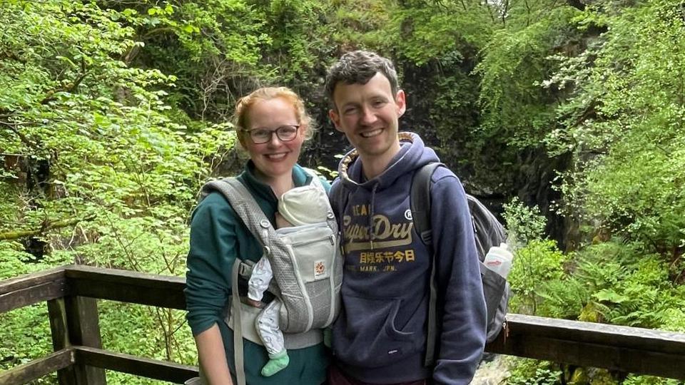 Lucy and Daniel Slavin stand with woodland seen behind them. Lucy is carrying their young baby in a baby carrier on her chest.