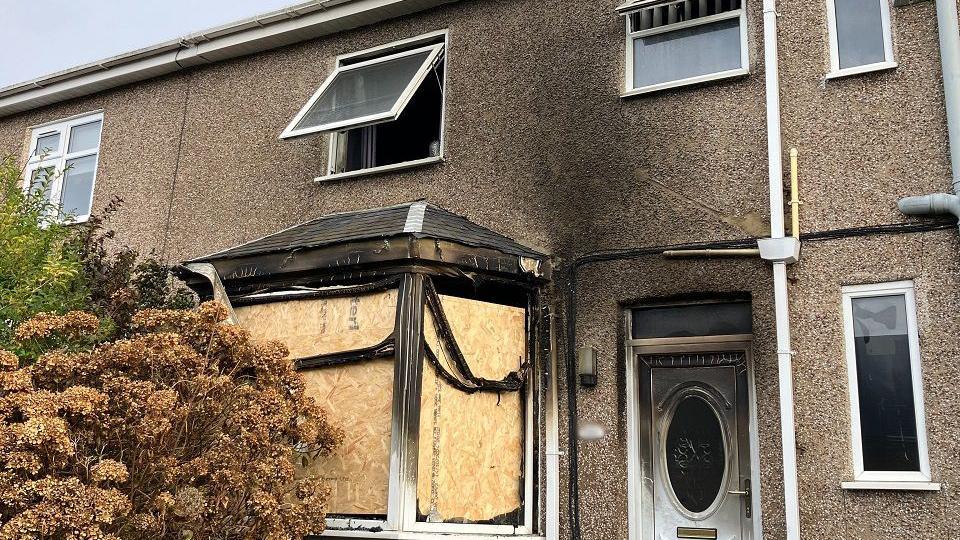 A brown, pebble-dashed, semi-detached house blackened by smoke. The white front door is covered in smoke damage and the front bay window is boarded up.