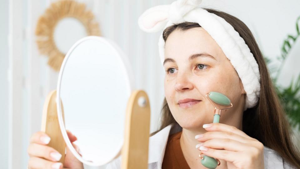 A young woman with dark hair uses a small roller on her cheek while looking into a mirror