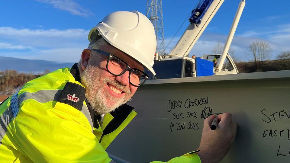 Derry Crorken, wearing glasses, a white hard hat and yellow high-vis jacket, signs one of the steels for the new building. He has a short white beard and is smiling at the camera