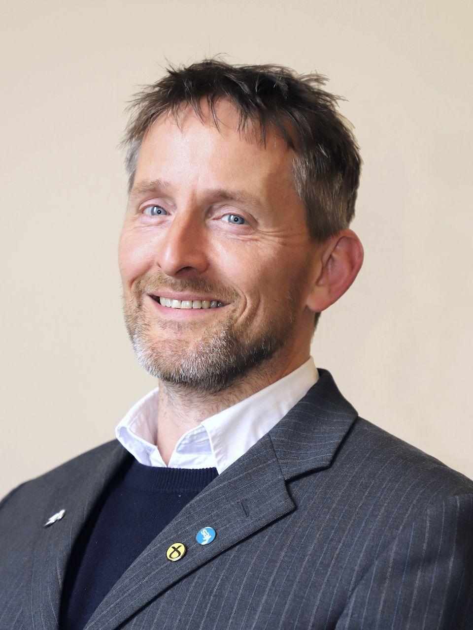 A man with dark hair and a beard in a suit jacket with a jumper and shirt underneath