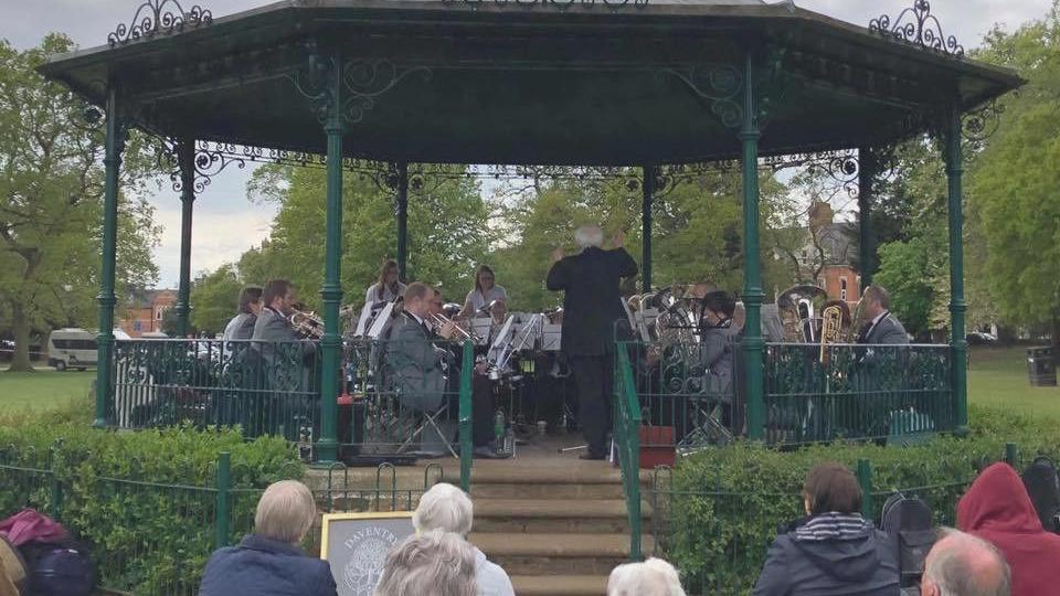 The bandstand with green poles and railings and a black roof. There is a band dressed in white shirts and dark trousers playing various brass and percussion instruments. There is a low hedge in front of the bandstand and stone steps leading down from the raised platform to the ground. A number of people are standing in front of the bandstand listening to the music.