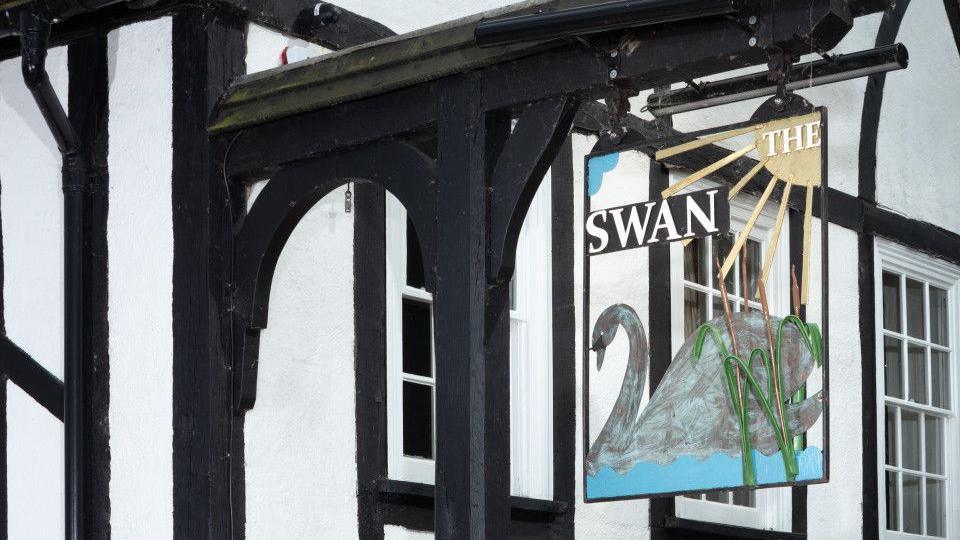 The white painted exterior of The Swan with black painted beams. A sign of a swan with writing which says THE SWAN in white lettering, hangs from the outside of the building.