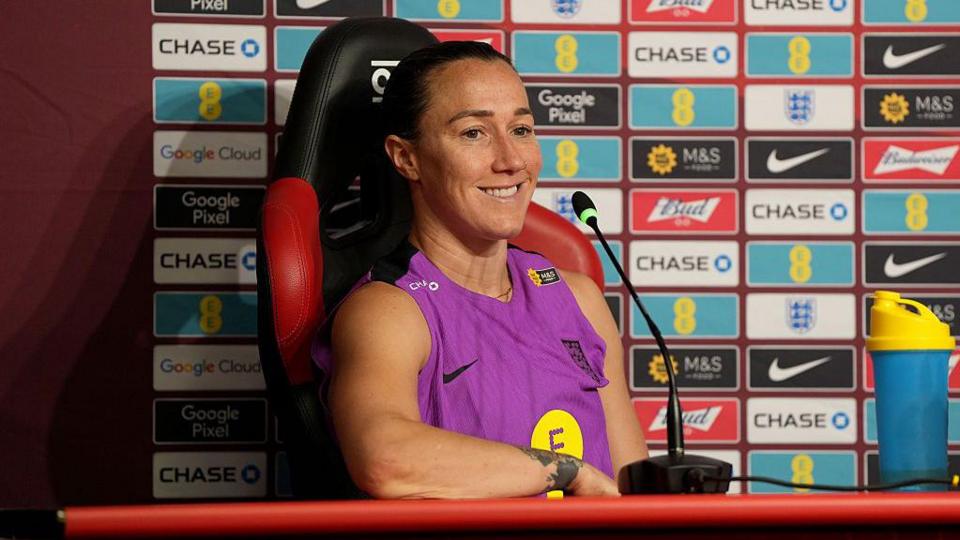 Lucy Bronze, wearing an England training vest, smiles as she speaks at a press conference