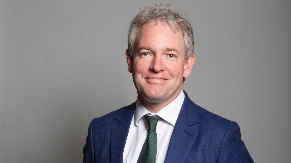 Danny Kruger wearing a white shirt, blue suit and green patterned tie. He has grey hair and blue eyes and is standing against a grey background, smiling at the camera.