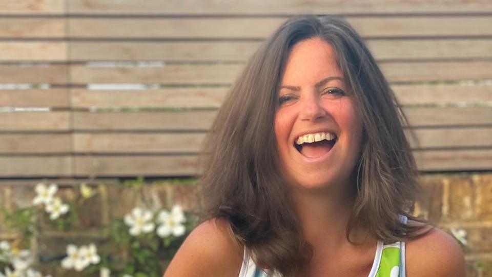 "Bowelbabe" Dame Deborah James smiles to camera. She looks happy and has bobbed brunette hair which is sitting on her shoulders. She is stood in front of a garden wall and fence on a bright sunny day. She is wearing a fundraising vest. 