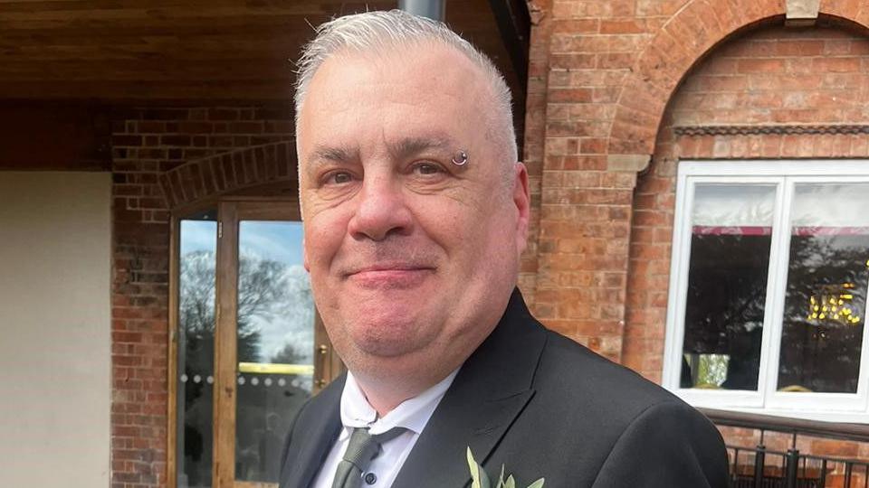 Sean is short cropped grey hair and a ring piercing on the end of the right eyebrow. He is wearing a black blazer over a white shirt and grey tie. There is a flower in the right breast pocket. He is standing in front of a redbrick building and smiling into the camera.