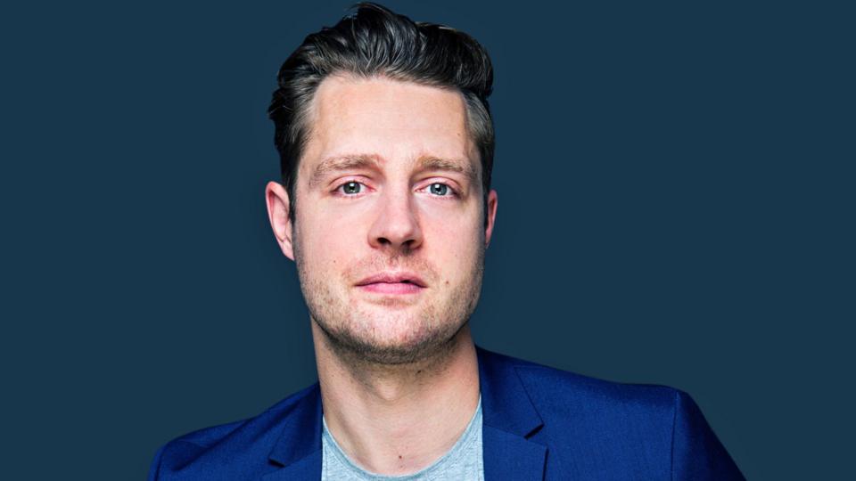 Comedian Max Dickins, wearing a blue suit jacket and light grey t-shirt, fills the frame and looks directly at the camera. He is stood in front of a dark blue background