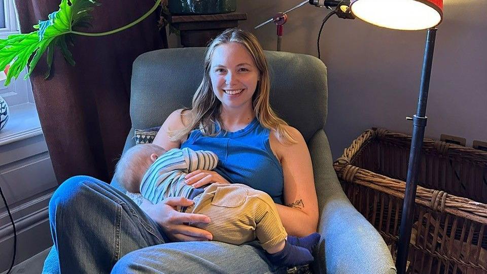 A woman with shoulder length blonde hair and wearing jeans and a blue vest top is sitting on a chair with her baby. She's in a house, with a window behind her and a lamp beside her