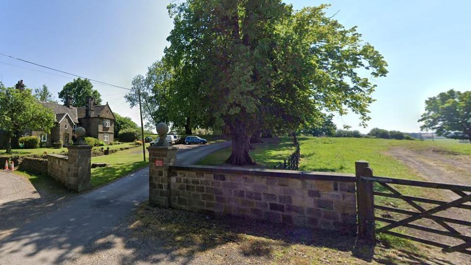 A driveway, lined with trees and with grass area either side of it can be seen from the road. There is a gate as well as a small wall which opens onto the driveway.