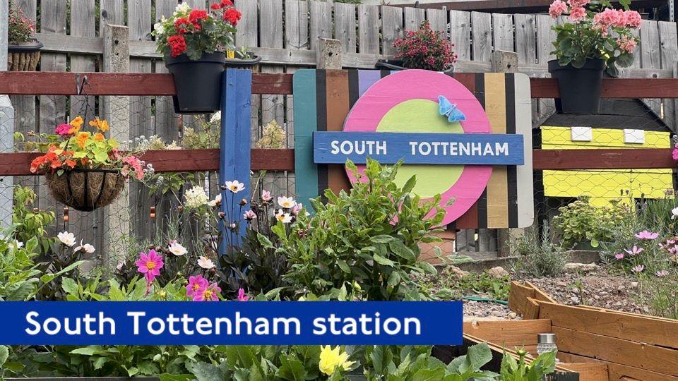 In the background, the South Tottenham roundel is in pink and yellow and it has a large, blue butterfly on it.  In the foreground there are lots of colourful flowers and hanging baskets