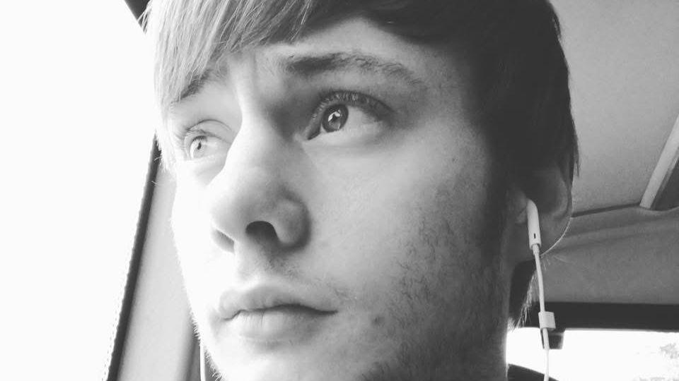 A black-and-white photo of Joe Nihill, a 23-year-old man. He is white with a beard, is looking slightly off camera, seemingly out of the window of a bus. He has earphones in. It is a close-up selfie.