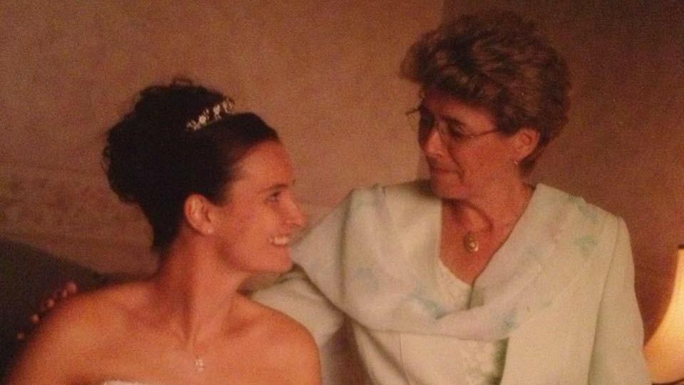 Simone Sains is pictured with her mother on her wedding day. The two women are smiling at each other. Ms Sains' mother has her arm around her daughter and her hand on her shoulder. They appear to be sitting on a sofa in a living room. There is a lamp beside them.