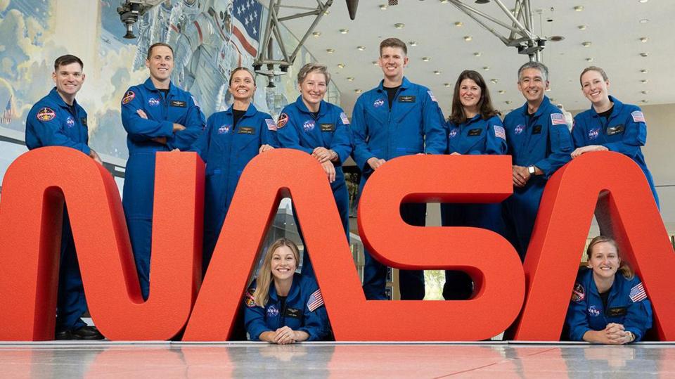 Ten new astronauts all in blue nasa overalls behind a red Nasa sign