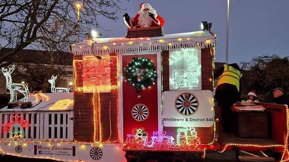 Red and white Santa's float with Christmas-themed décor including a white reindeer and a green wreath and a Santa on top of it