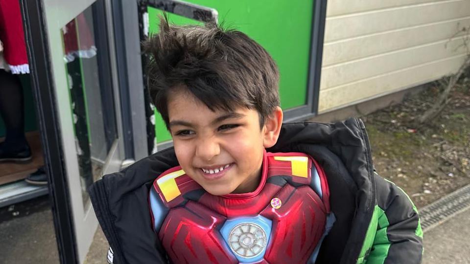 Saleh Aslam smiling. He is outside a school building and wearing a coat and superhero outfit. He has dark hair and is looking to his right as he smiles. 