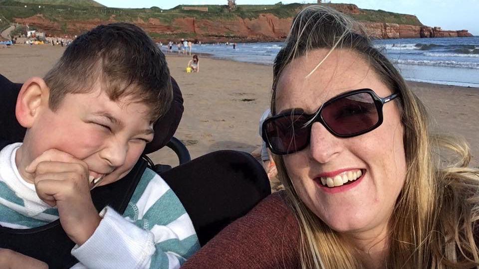 Brody on the left, wearing a striped top with Nikki on the right, wearing sunglasses. They are at the beach and you can see sand and sea behind them. Brody is in a wheelchair and they are both smiling