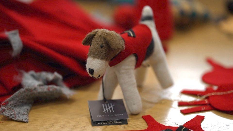 A little white and brown felt dog wearing a red jacket stands on a table among offcuts of other bits of fabric