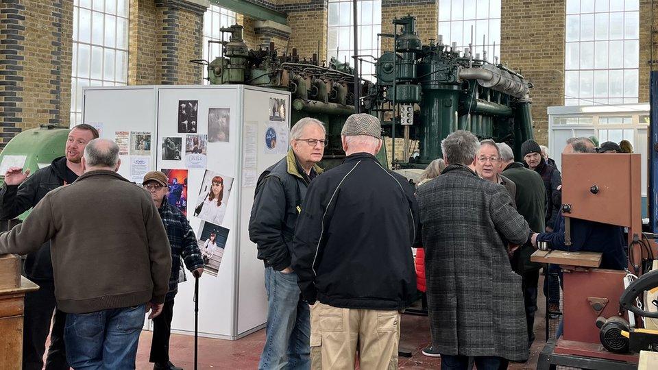 Small groups of men engage in conversation in front of two ten foot water pump engines