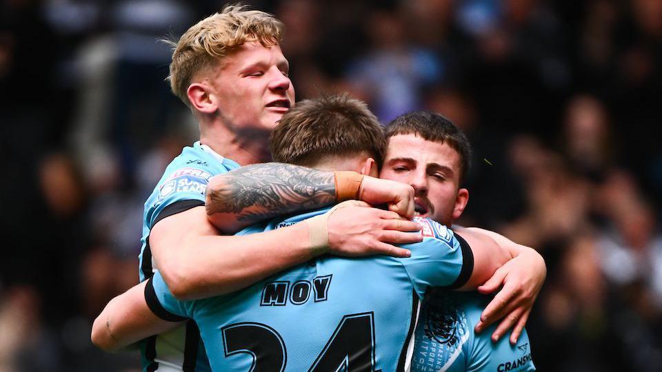 Hull FC players Lloyd Kemp (left) and Amir Bourouh (right) hug try-scorer Logan Moy, all players dressed in light blue.