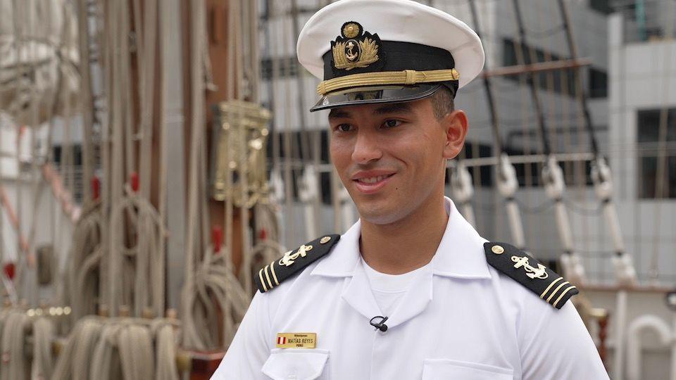 A smiling young naval cadet wears a smart white shirt with black epaulettes on the shoulders that have gold anchors embroidered on them. He wears a white military cap with black peak and gold embellishment. There is rope and rigging in the background