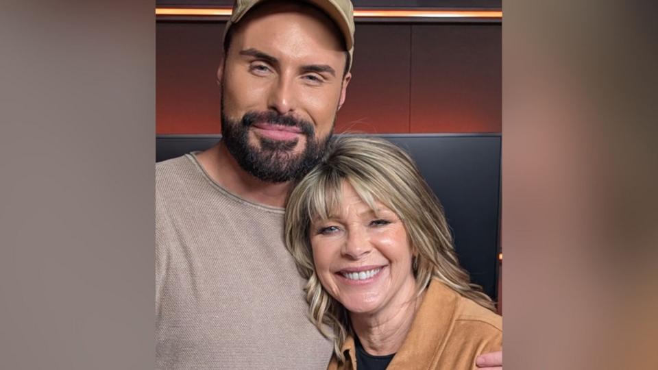Ruth Langsford stands next to Rylan, both are smiling towards the camera.