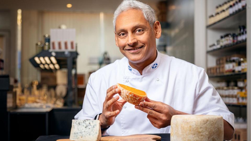 A man in chefs whites with grey hair holds up a chunk of cheese with a couple of other blocks of cheese in front of him and what looks like a blurred kitchen area behind him