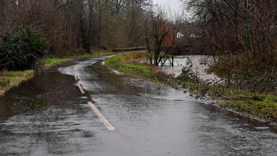 A flooded country road with standing water and nearby river overflowing into surrounding trees.