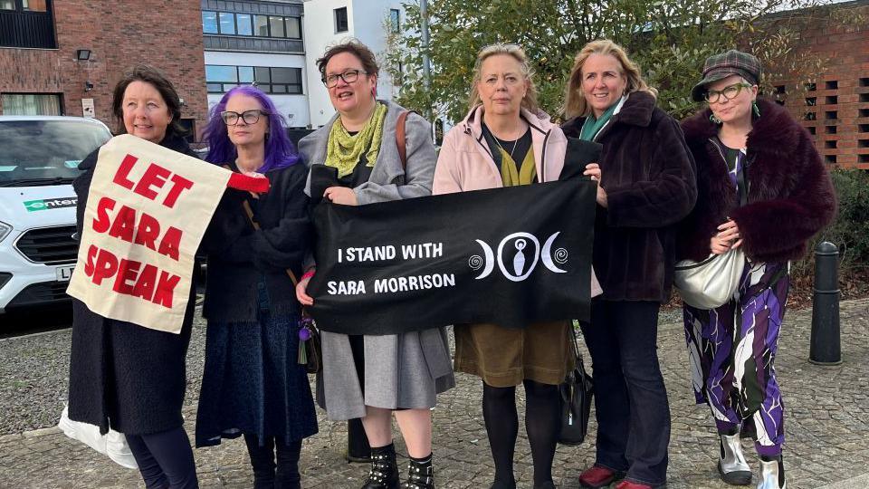 A group of women standing next to each other. There are six of them wearing a range of different coloured clothing. From left to right a woman with long brown hair, a lady with long purple hair and dark glasses. a women with short brown hair and square glasses, then there is a woman with long blonde hair and next to her is a women with blonde hair and a lady wearing a hat and green glasses.