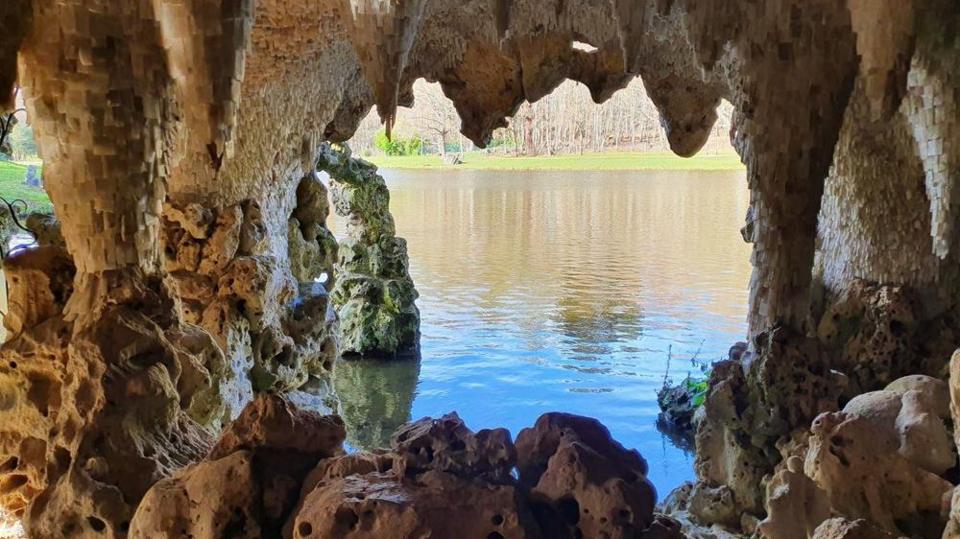 An interior view from inside a grotto, framed by rough stone openings, looks out onto a lake, with light reflecting on the water.