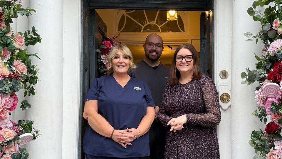 Three people - Barbara Pointon with son Dan, another practitioner, and Labour MP Leigh Ingham - are standing in a doorway with foliage either side. There are two women in the front, one wearing a blue top and the other a grey and brown dress. A man is standing behind them wearing a grey top.