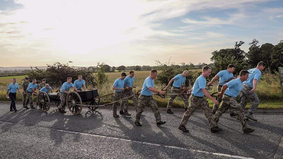 Volunteers from the charity pulling the gun on an uphill road in 2019. Some are walking alongside and behind it, pushing it forward. Most walk in front, pulling ropes which are chained to the cannon's wheels. 