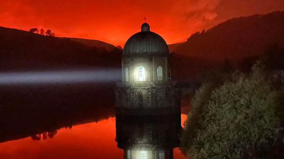 A lighthouse is pictured in the water, shining a light. Above it is a red sky, lit up by wildfires in the distance. The red colour is also reflected in the water.
