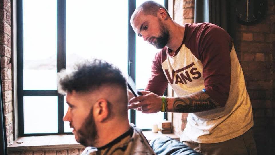 A man with bald head, beard and arm tattoos cutting a man's hair 